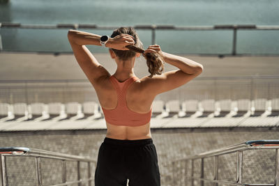 Back view of unrecognizable slim female athlete in sportswear standing putting on hairband on urban embankment near river and relaxing after exercising in summer day