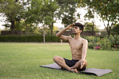 Full length of shirtless man sitting on grass against trees