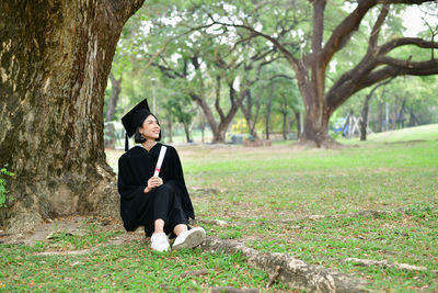 Student wearing graduation gown while sitting on field at park