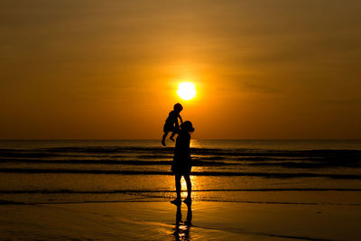 Silhouette mother holding son while standing on shore against sea during sunset