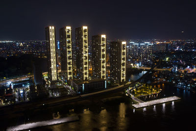 Illuminated modern buildings by river against sky at night