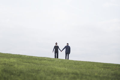 Rear view of couple holding hands standing on grassy land against sky