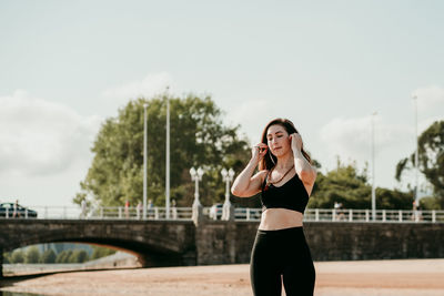 Calm fit female in sports bra and leggings standing on beach and preparing for training while touching hair