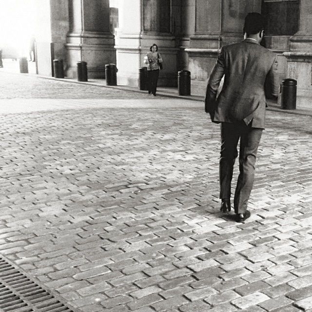 REAR VIEW OF A MAN WALKING ON COBBLESTONE STREET