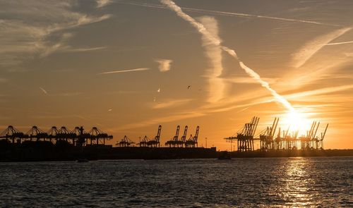 Silhouette cranes at commercial dock against sky during sunset