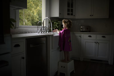 Side view of girl wearing pink cape washing hands in kitchen sink while standing on stool at home