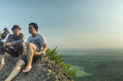 Young men looking away sitting on rocks against sky