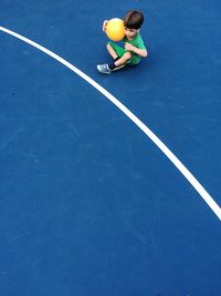 High angle view of boy with ball sitting at tennis court