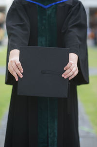 Midsection of student wearing graduation gown while holding mortarboard outdoors