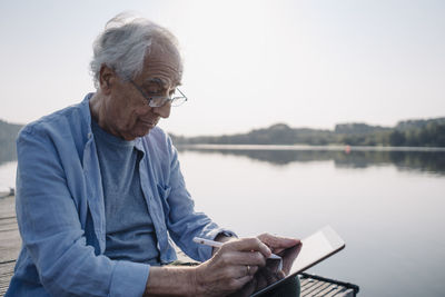 Senior man wearing eyeglasses using digital tablet while sitting on pier