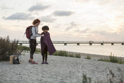 Woman wrapping girl with towel at beach against sky during sunset