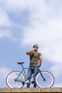 Low angle view of mid adult man talking on mobile phone while standing with bicycle against sky