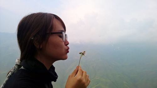 Close-up of young woman blowing dandelion seeds against mountains