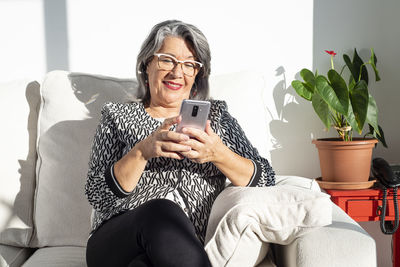 Smiling old lady sitting on a sofa and using a smartphone mobile