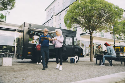 Senior male and female customers with street food walking against commercial land vehicle in city