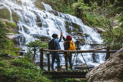 Friends with camera giving high-five by waterfall in forest