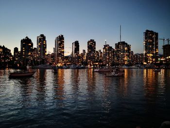 Sailboats in river by illuminated buildings against clear sky