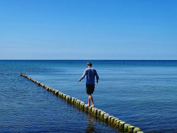 Rear view of man walking on wooden posts over sea against blue sky