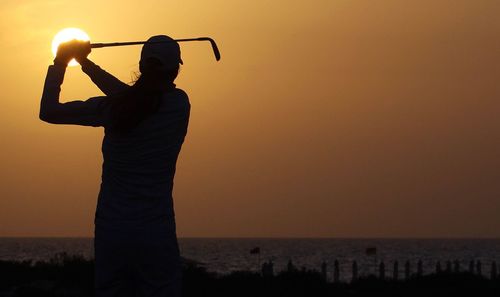 Silhouette woman with golf club standing at beach against sky during sunset