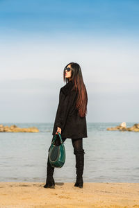 Portrait of young woman standing at beach against sky