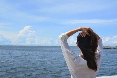 Rear view of woman with hand in hair standing by sea against sky