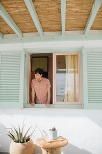 Emotionless young male in casual shirt looking out window while standing in contemporary house on sunny weather