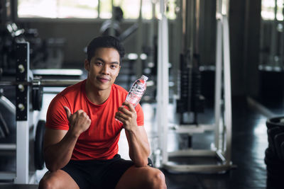 Portrait of young man showing thumbs while sitting with water bottle in gym