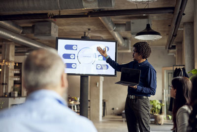 Businessman holding laptop while gesturing at television set during meeting in office