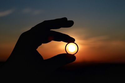 Silhouette of human hand holding ring against sky during sunset