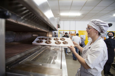 Side view of female baker in uniform with tray of bread dough turning on oven while preparing for baking in bakery school with people