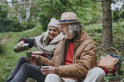 Smiling woman pouring water from thermos by senior man in forest