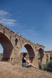 Woman traveler explorer in a black hat sit next to the destroyed old stone bridge in the summer