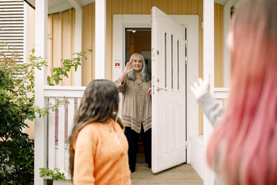 Senior woman waving hand at granddaughter from entrance of house
