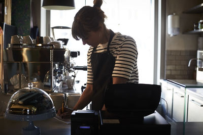 Barista writing checklist while standing at checkout counter in cafe