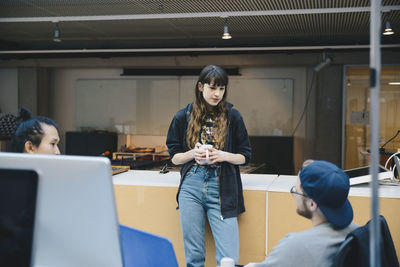 Female computer programmer holding coffee mug while discussing with colleagues in office