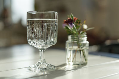 Glass of mineral water and small bouquet of flowers in jar on table in sunlight, healthy lifestyle