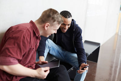 Smiling man showing mobile phone to friend while sitting on bench in university