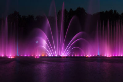 Illuminated fountain by river against sky at night