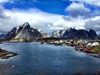 Houses on riverbank by mountains against cloudy sky