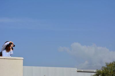 Woman photographing while standing on terrace against blue sky