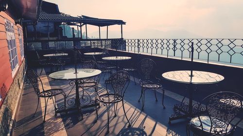 Empty chairs and tables by sea against sky during sunset