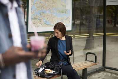 Businesswoman eating sandwich while sitting at bus stop in city