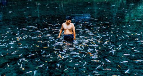 Shirtless young man standing amidst fish in lake