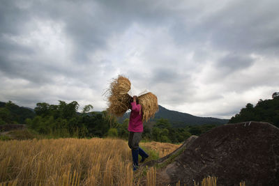 Man carrying hay bale while walking on field against cloudy sky