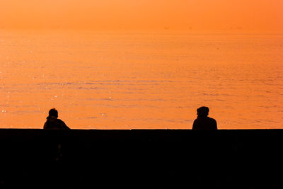 Silhouette people sitting on land against orange sky