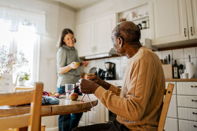 Side view of senior man knitting while talking with female nurse in kitchen at home