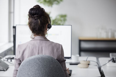 Rear view of businesswoman using headset in office in front of computer screen