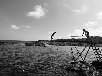 Woman on diving platform with man jumping in sea against sky