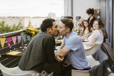 Affectionate gay couple kissing while sitting with friends at dinner party in balcony