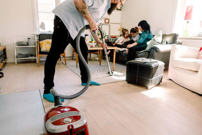 Man cleaning floor with vacuum cleaner while family sitting on sofa